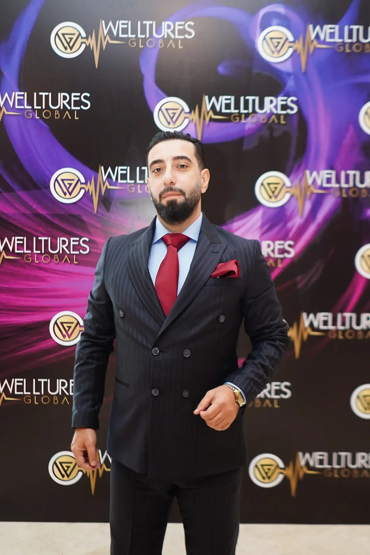 ببدلة كحلية مقلّمة وربطة عنق حمراء أمام جدار Welltures Global الصحفي